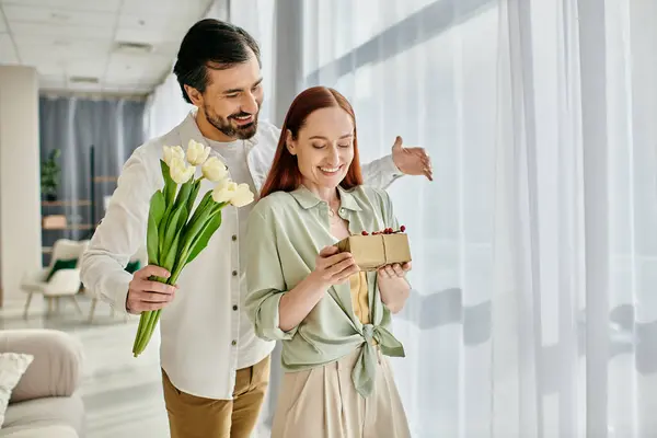 A beautiful adult couple, a redhead woman and a bearded man, hold a gift box in front of a window in a modern apartment. — Stock Photo