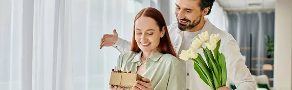 A bearded man holds a bouquet of flowers in front of a window, creating a romantic atmosphere in their modern apartment. — Stock Photo