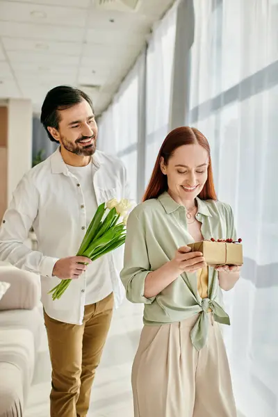 Un homme barbu et une rousse marchent dans un salon moderne, tenant une boîte cadeau. — Photo de stock