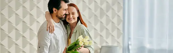 A redhead woman and bearded man embrace lovingly in front of a chic wall in their modern apartment. — Stock Photo