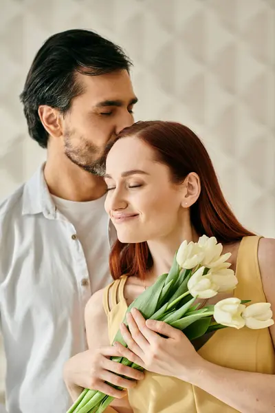A redhead woman and bearded man embrace and kiss while holding a bouquet of tulips in a modern apartment. — Stock Photo