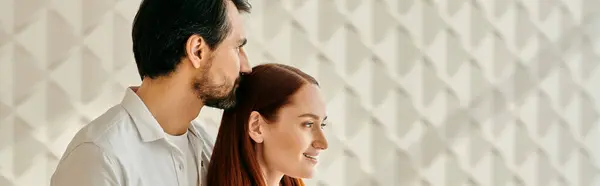 A beautiful adult couple, a redhead woman and a bearded man, embracing in front of a white wall in their modern apartment. — Stock Photo