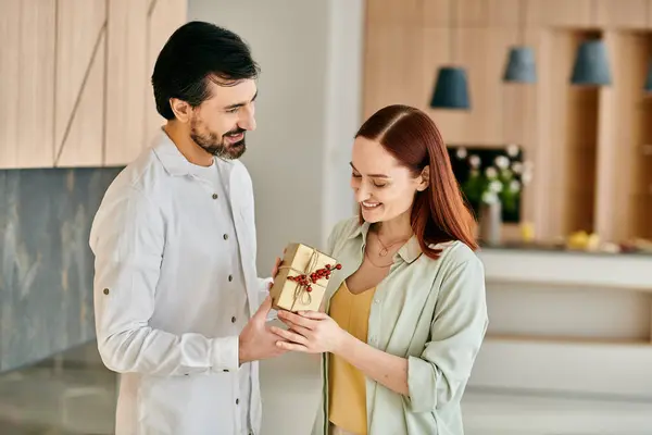 Una donna dai capelli rossi e un uomo barbuto condividono una scatola regalo in una cucina moderna, scambiandosi sorrisi e diffondendo gioia. — Foto stock