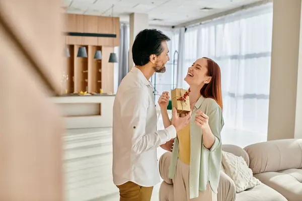 Uma mulher ruiva e um homem barbudo estão em pé em uma sala de estar moderna, alegremente segurando um presente entre eles. — Fotografia de Stock