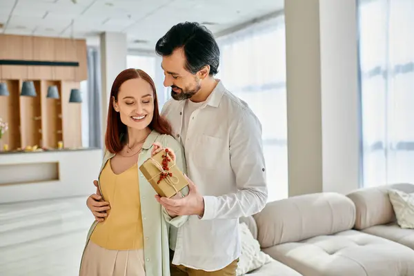 Una bella coppia di adulti, una donna rossa e un uomo barbuto, con una scatola regalo nel loro moderno soggiorno. — Foto stock