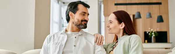 A beautiful adult couple, a redhead woman and a bearded man, sitting on a couch, gazing lovingly at each other in a modern apartment. — Stock Photo