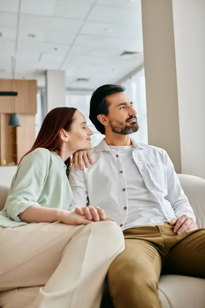 Una hermosa pareja de adultos, una mujer pelirroja y un hombre barbudo, se sientan en un sofá en una sala de estar moderna, pasando tiempo de calidad juntos. - foto de stock