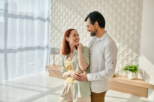 A beautiful adult couple, a redhead woman, and a bearded man, hugging affectionately in a modern living room. — Stock Photo