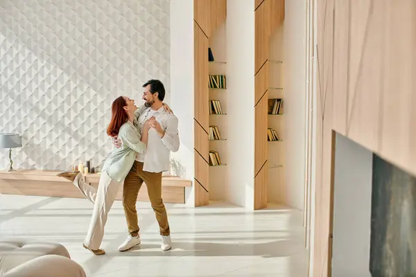 A beautiful adult couple, a redhead woman and a bearded man, dancing joyfully in their modern living room. — Stock Photo