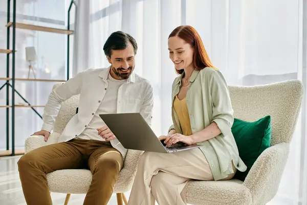 Una donna rossa e un uomo barbuto siedono su una sedia, concentrati su uno schermo del computer portatile in un appartamento moderno, nel profondo della conversazione. — Foto stock