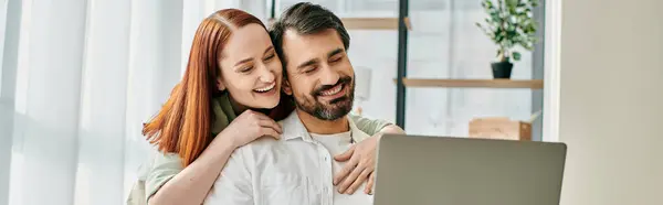 A redheaded woman and a bearded man hugging while enjoying a moment together by looking at a laptop in a modern apartment. — Stock Photo