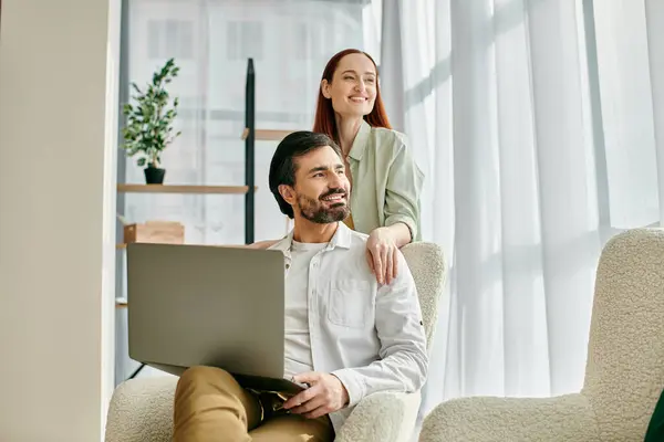 Dicht an dicht sitzen eine rothaarige Frau und ein bärtiger Mann in einem Stuhl, vertieft in die gemeinsame Nutzung eines Laptops in ihrer modernen Wohnung.. — Stockfoto