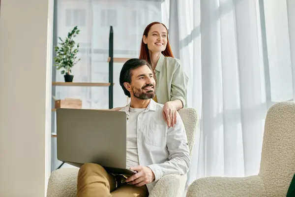 A redhead woman and bearded man sit closely in a chair, engrossed in using a laptop together in their modern apartment. — Stock Photo
