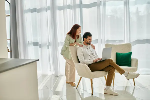 A redhead woman and bearded man sit in a chair, using a laptop together in a modern apartment, enjoying quality time. — Stock Photo