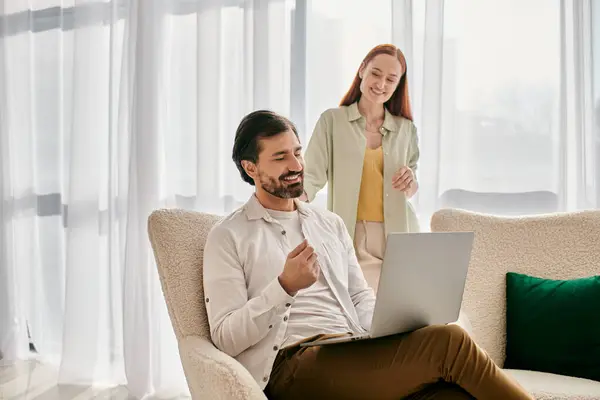 Eine rothaarige Frau und ein bärtiger Mann sitzen auf einer Couch und benutzen gemeinsam einen Laptop in einer modernen Wohnung. — Stockfoto