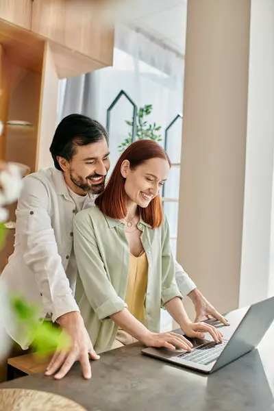 Una mujer pelirroja y un hombre barbudo absortos en la pantalla de un ordenador portátil, mientras que de pie en su cocina moderna. - foto de stock