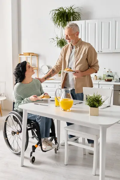 Um homem em uma cadeira de rodas serve comida para sua esposa deficiente em sua cozinha em casa. — Fotografia de Stock