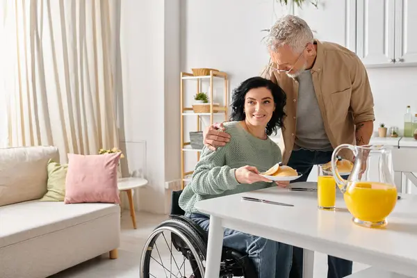A man in a wheelchair lovingly feeds his disabled wife a piece of food in their home kitchen. - foto de stock