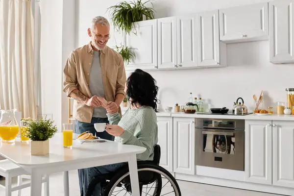 A husband stands next to his disabled wife in a wheelchair in the cozy kitchen of their home, sharing a moment of togetherness. — Photo de stock
