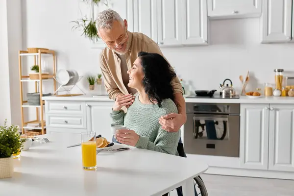 Un marito sta accanto alla moglie disabile in sedia a rotelle, condividendo un momento nella loro accogliente cucina a casa. — Foto stock