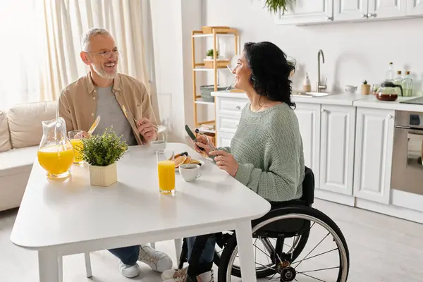 Un hombre en silla de ruedas y una mujer en silla de ruedas hablando en la cocina en casa. - foto de stock