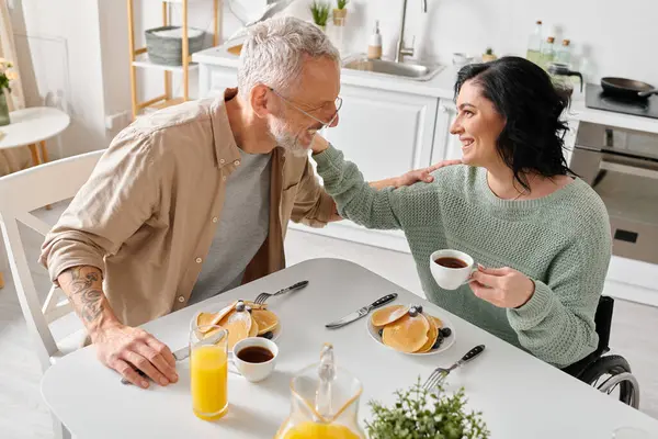 Une femme handicapée dans un fauteuil roulant et son mari apprécient volontiers le petit déjeuner ensemble à une table dans leur cuisine confortable. — Photo de stock