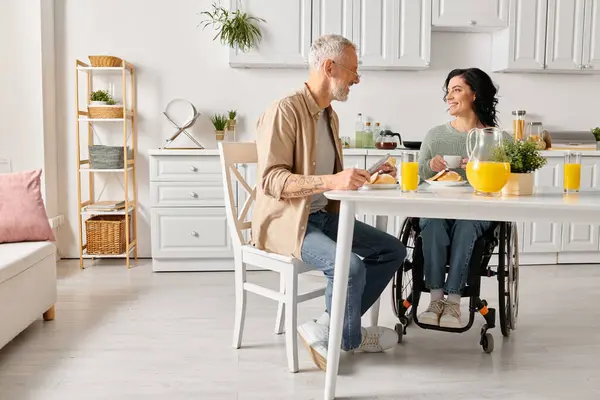 Una mujer discapacitada en silla de ruedas y su marido compartiendo un momento tranquilo en la mesa de su cocina en casa. - foto de stock