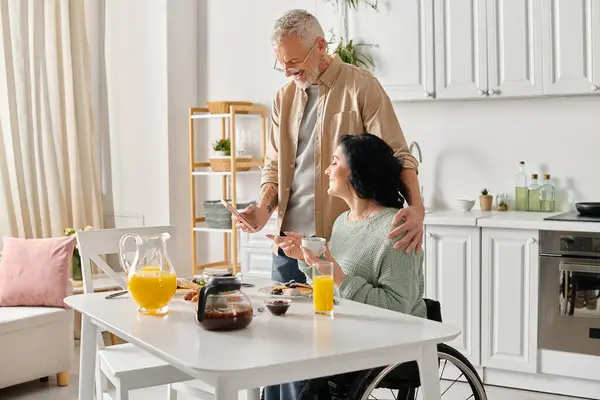Un marito sta accanto alla moglie disabile su una sedia a rotelle, offrendo sostegno e compagnia nella loro cucina di casa.. — Foto stock
