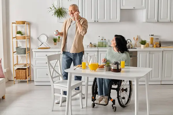 A man stands beside his disabled wife, who is sitting in a wheelchair at a table in their kitchen at home. — стокове фото
