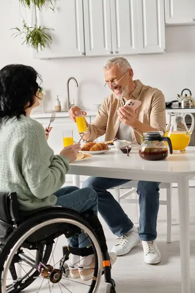 Un hombre y una mujer en silla de ruedas comparten un acogedor desayuno en una cocina en casa. - foto de stock