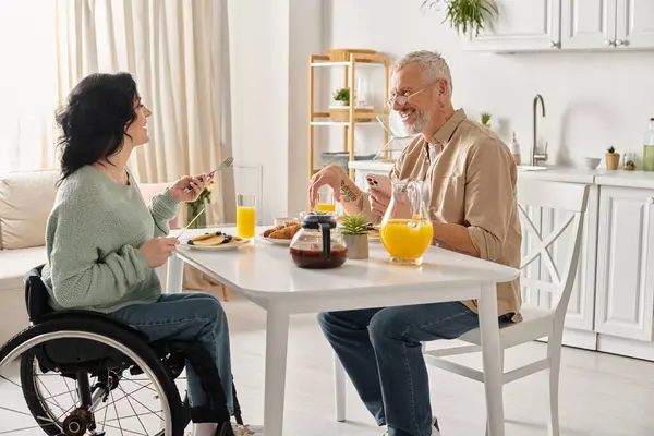 Une femme handicapée en fauteuil roulant et son mari s'assoient à une table, prenant le petit déjeuner dans leur cuisine à la maison. — Photo de stock
