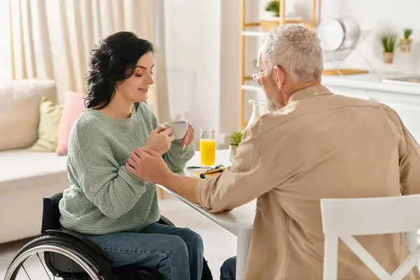 A woman in a wheelchair graciously hands a cup of coffee near man in a kitchen at home. — Stock Photo