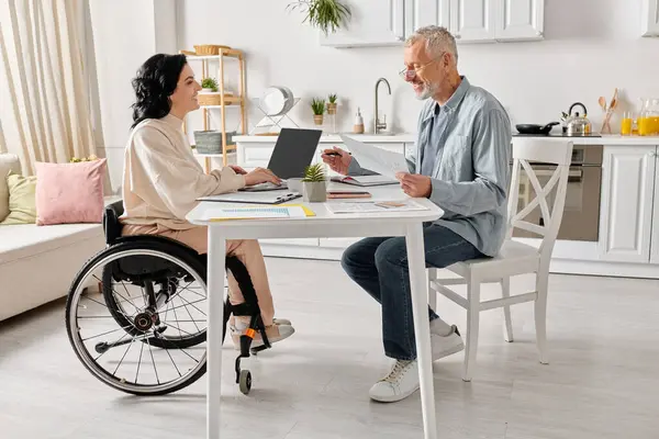 A man and a woman in wheelchair sit together at a table in their kitchen at home. — Stock Photo