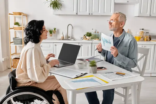 Una mujer discapacitada en silla de ruedas se sienta junto a su marido en una mesa de cocina, revisando los papeles juntos. - foto de stock