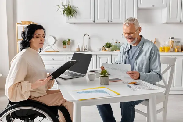 A man sits at a table with his disabled wife in a wheelchair, both focused on a laptop screen in their cozy kitchen at home. — Stock Photo
