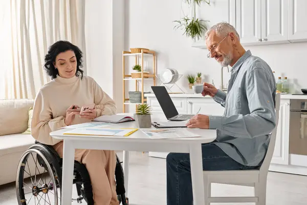 Une femme en fauteuil roulant et un homme avec un ordinateur portable, partageant un moment de convivialité dans leur cuisine à la maison. — Photo de stock