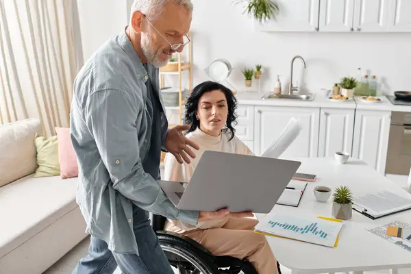 Uma mulher em uma cadeira de rodas usando um computador portátil em um ambiente acolhedor em casa, perto do marido — Fotografia de Stock