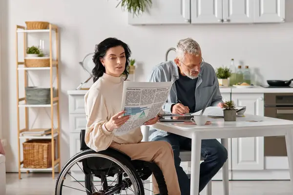 A man in a wheelchair reads a newspaper while his wife sits at a table in their kitchen at home. - foto de stock