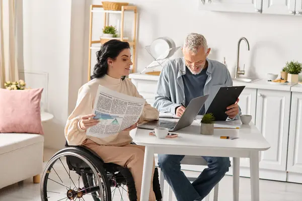 A man and disabled woman sit at a table, focusing intently on a laptop screen in their cozy kitchen at home. - foto de stock