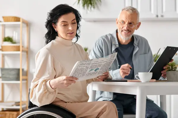 A husband and his disabled wife, seated in the kitchen, reding newspaper — Stock Photo