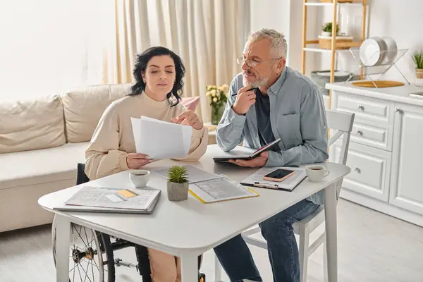 Ein Mann und eine behinderte Frau im Rollstuhl sitzen zu Hause an einem Tisch in ihrer Küche und planen ihr Budget — Stockfoto
