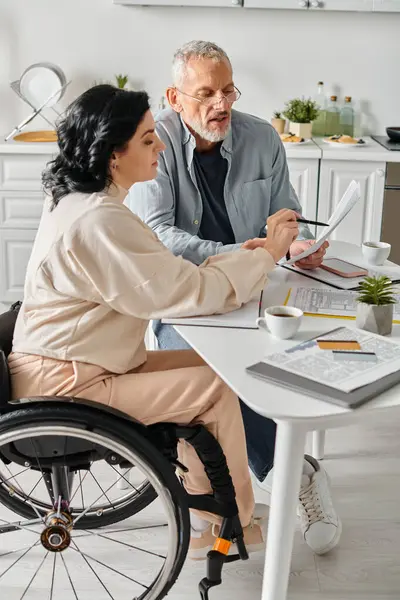 A disabled woman in a wheelchair and her husband comfortably seated at a table in their kitchen at home. - foto de stock