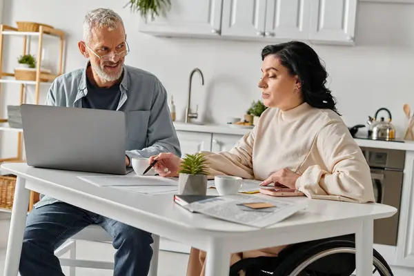 Un homme et une femme, la femme en fauteuil roulant, assis ensemble à une table avec un ordinateur portable, absorbé dans ce qui est à l'écran. — Photo de stock
