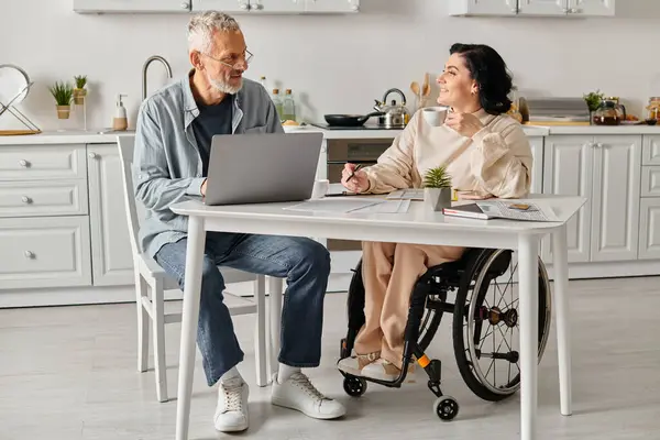 A husband and his disabled wife sit at a kitchen table working on a laptop together. — Stock Photo