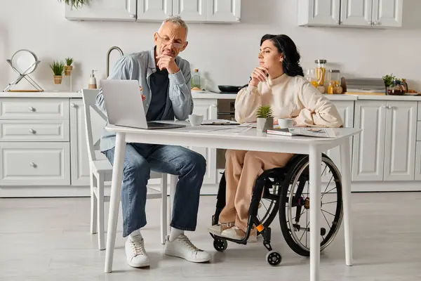 A woman in a wheelchair and her husband sit at a table, engaged with a laptop in a cozy kitchen setting at home. — Stock Photo