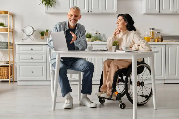 Um homem e uma mulher com deficiência em uma cadeira de rodas trabalham juntos em uma mesa com um laptop em sua cozinha acolhedora em casa. — Fotografia de Stock