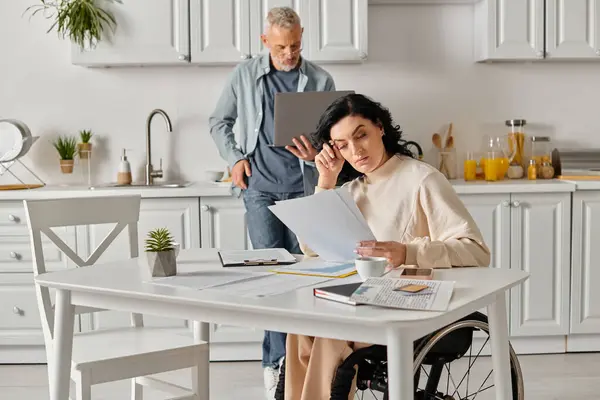 Eine behinderte Frau im Rollstuhl starrt zu Hause in der Küche auf ein Blatt Papier, ihren Mann an ihrer Seite.. — Stockfoto