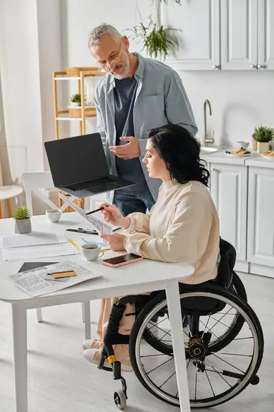 A man and a woman in wheelchairs absorbed in using a laptop in their kitchen at home. - foto de stock