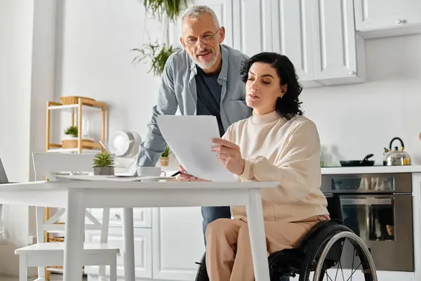 Un hombre en silla de ruedas y una mujer examinando un documento en una acogedora cocina en casa. - foto de stock