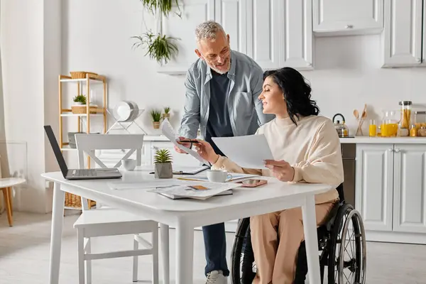 Un hombre y una mujer en sillas de ruedas están inspeccionando de cerca un pedazo de papel en su cocina en casa.. - foto de stock